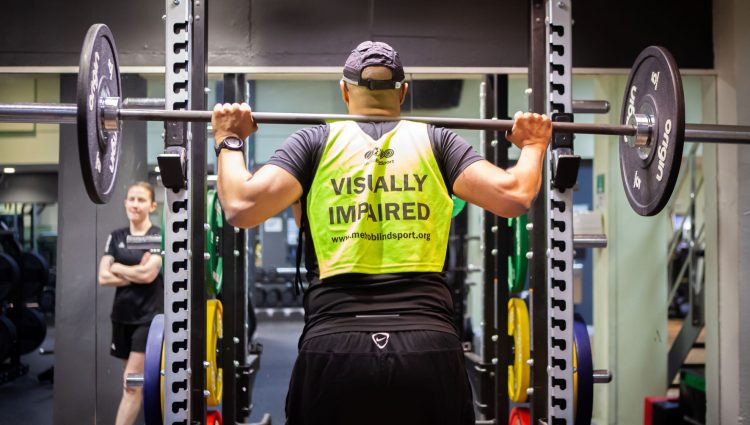 Image shows a gym goer in a squat rack at the gym. He has a barbell with weights on his back and is wearing a hi-vis vest which says 'Visually impaired'