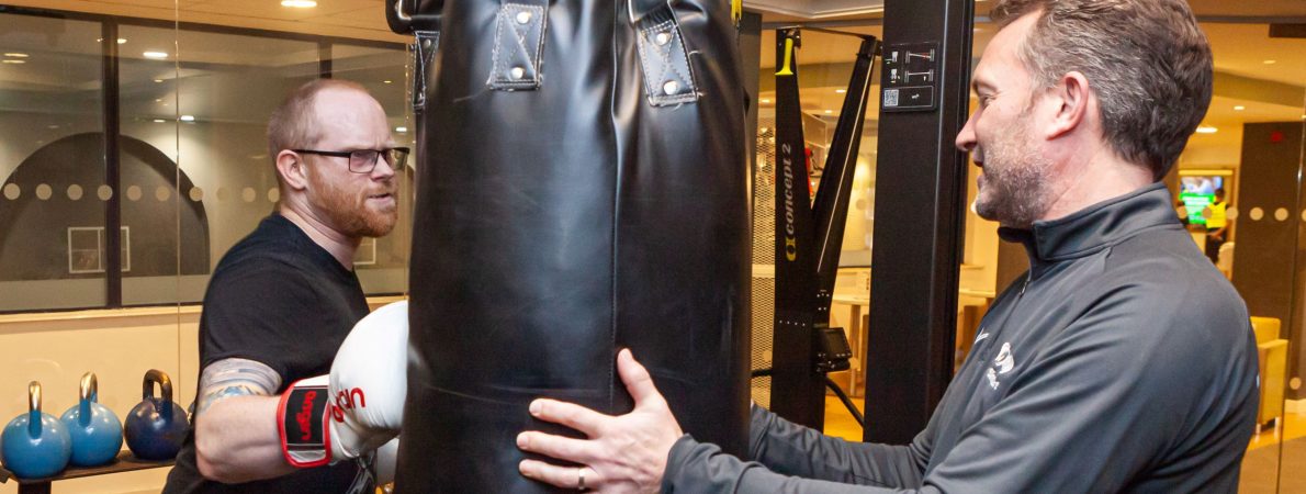 Photograph shows 2 men in a boxing gym. One is holding a punch bag and the other has boxing gloves on and is punching the punch bag.