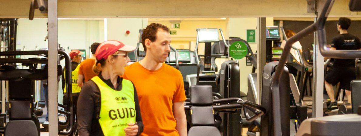 Photograph shows a woman guiding a visually impaired man through a gym. She is wearing a hi-vis vest on which says 'guide' on it. He is wearing a plain orange t-shirt.