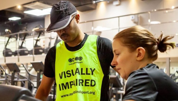 Photograph shows a man and a woman in a gym. He is wearing a hi-vis vest on which says 'visually impaired', and she is showing him how to use a piece of gym equipment.