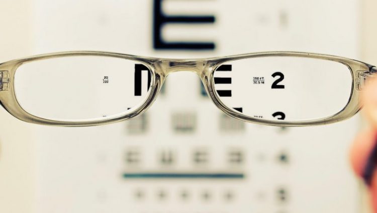 Photograph of an eye test chart being viewed through the aspect of a pair of sliver, metal rimmed glasses. the image through the lenses is clear, but otherwise blurred.