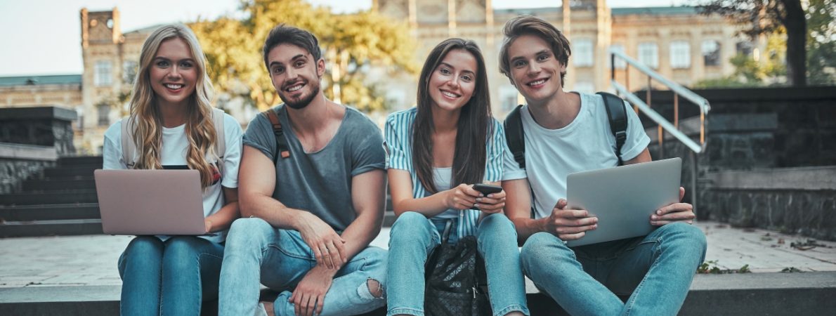 A group of two male and two female students, sat on stairs in front of a building with a yard, holding their laptops open and their phones, and smiling at the camera