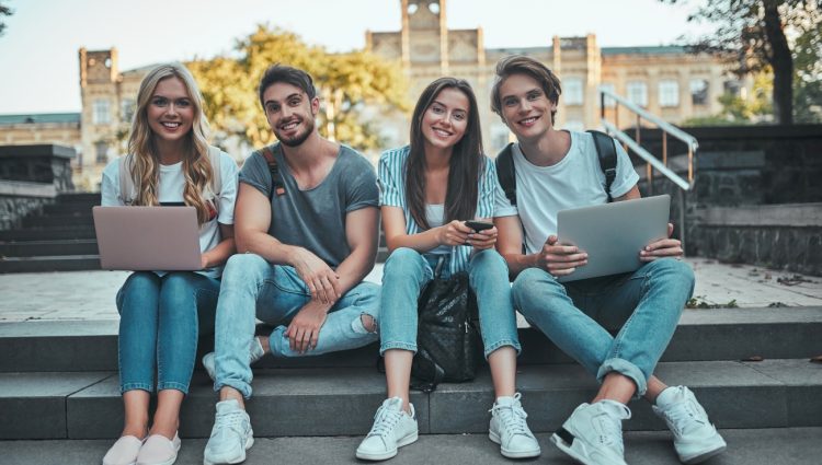 A group of two male and two female students, sat on stairs in front of a building with a yard, holding their laptops open and their phones, and smiling at the camera