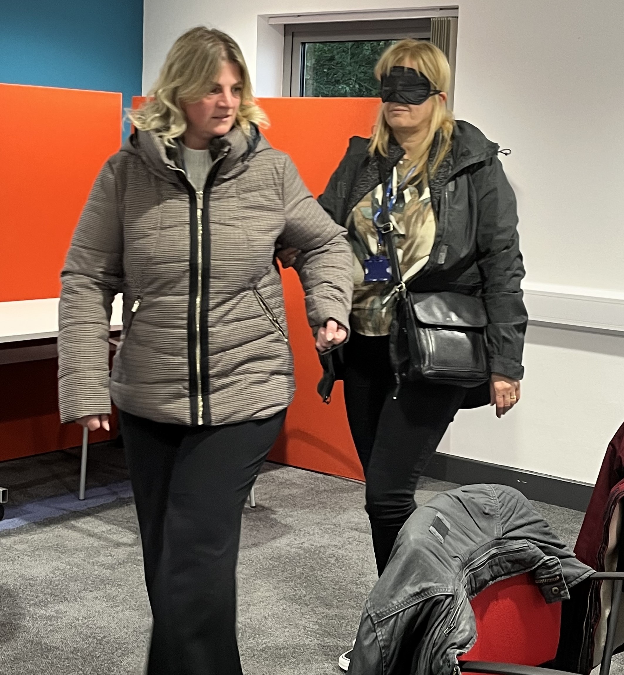 Photograph shows one woman wearing a blindfold, holding the hand of another woman who is guiding her