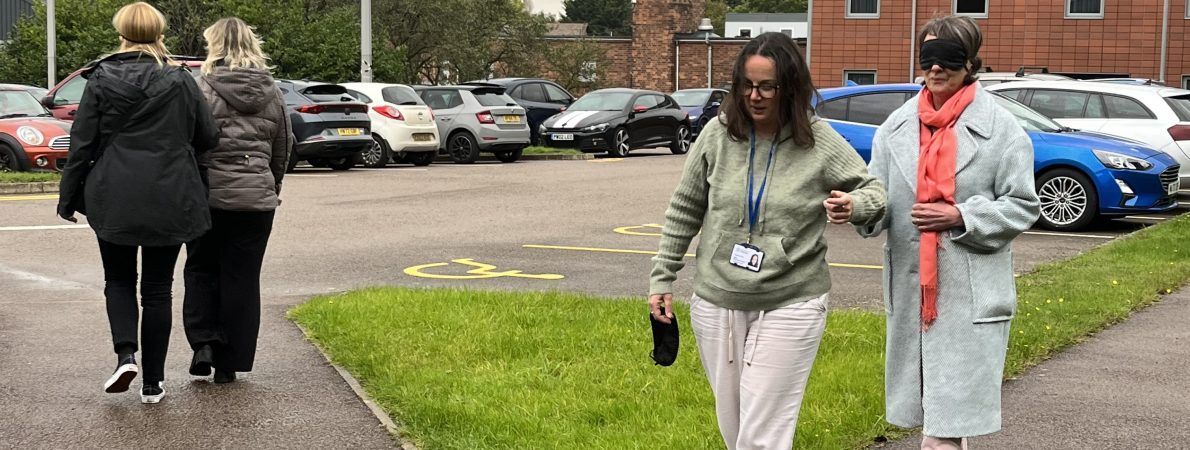 Photograph shows a pair of woman walking towards the camera in a car park. One woman is blindfolded and being guided by the other. There is another pair of people walking away from the camera.