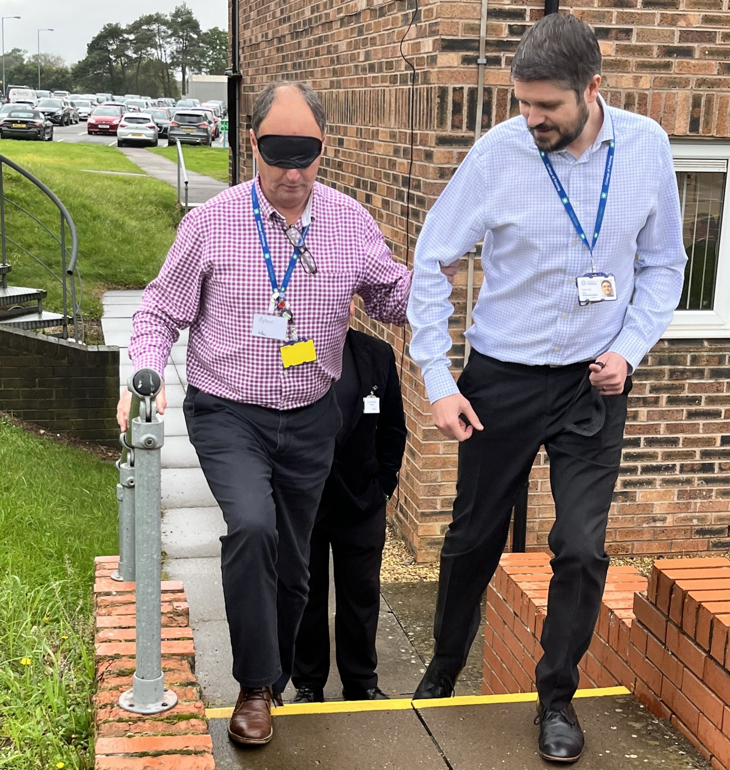 Photograph shows one man blindfolded, being guided up some steps by another man.