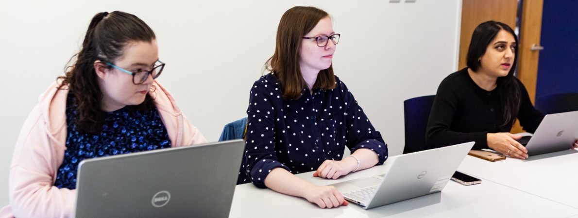 Three women sat at a big conference table, during a meeting. They are all have their laptops open.