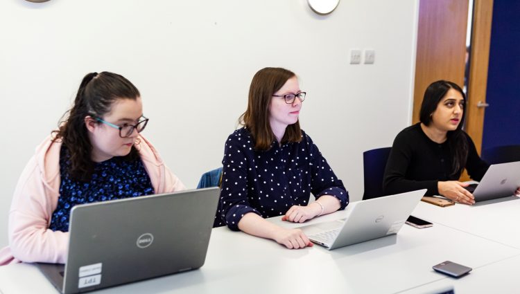 Three women sat at a big conference table, during a meeting. They are all have their laptops open.