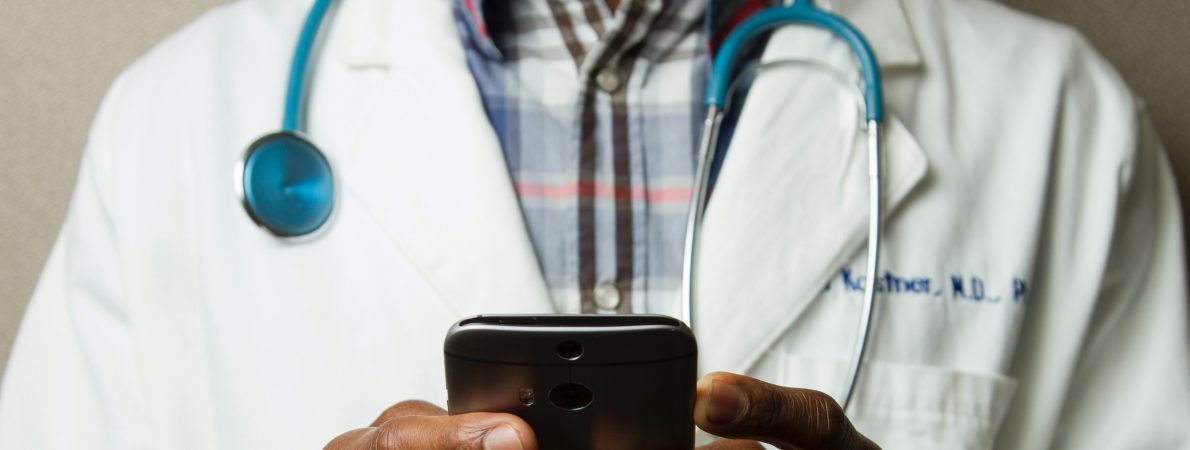 Photograph shows torso of a male doctor wearing a white coat with a stethoscope around his neck and holding a phone in both hands