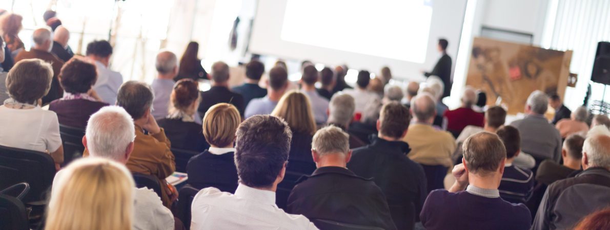 Photograph of an auditorium packed with people watching a presentation in the distance by a smartly dressed man standing in front of a screen. ages of the adult audience vary from young to old.