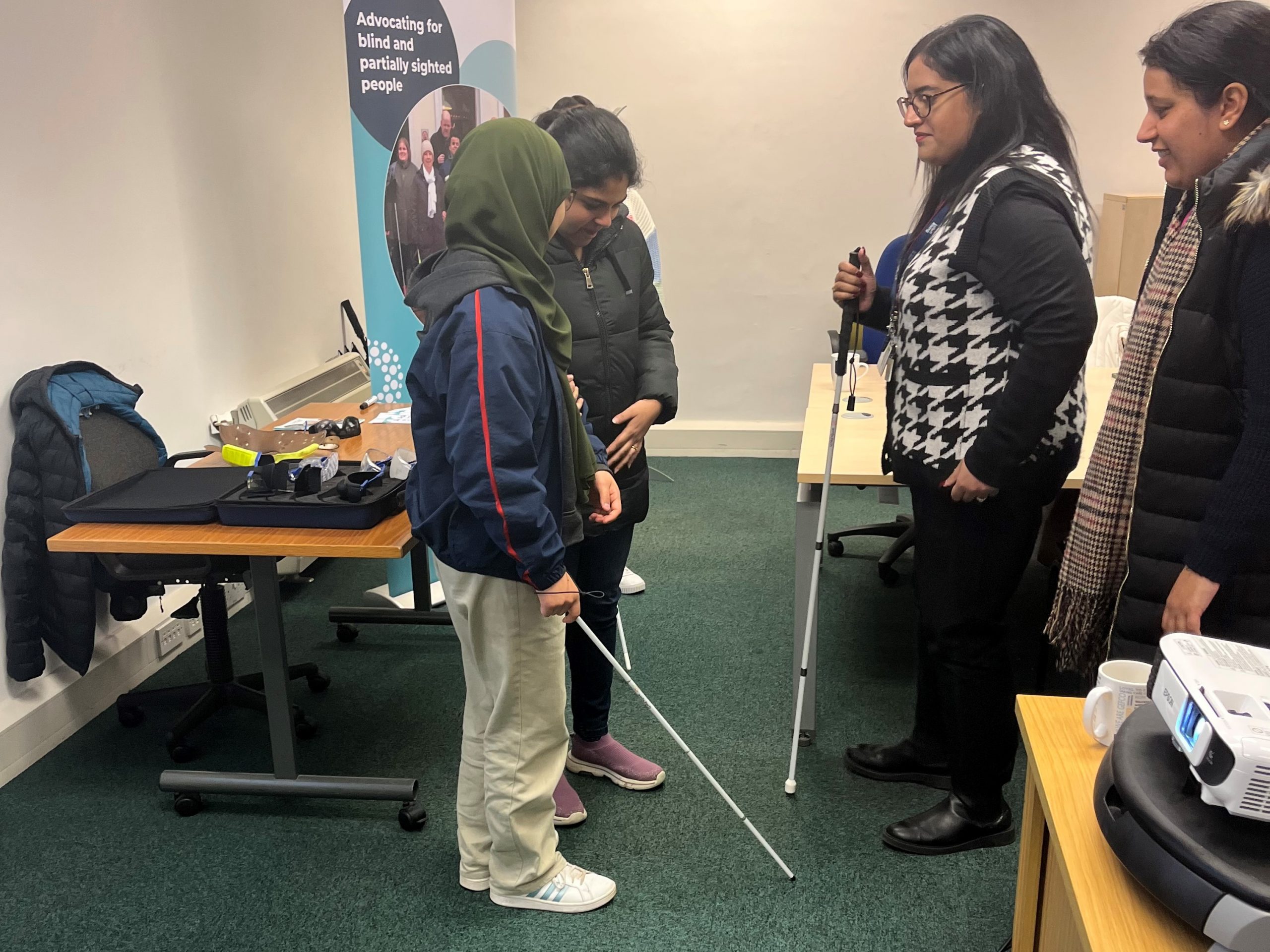 Photo shows Bedfordshire SLC members delivering a vision awareness session to Ethos Farm employees. There are 4 woman in camera shot. One is holding a white cane and showing another how to be a sighted guide. The other two are standing in front watching and listening.