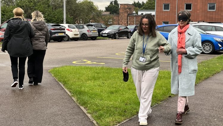 Photograph shows a pair of woman walking towards the camera in a car park. One woman is blindfolded and being guided by the other. There is another pair of people walking away from the camera.