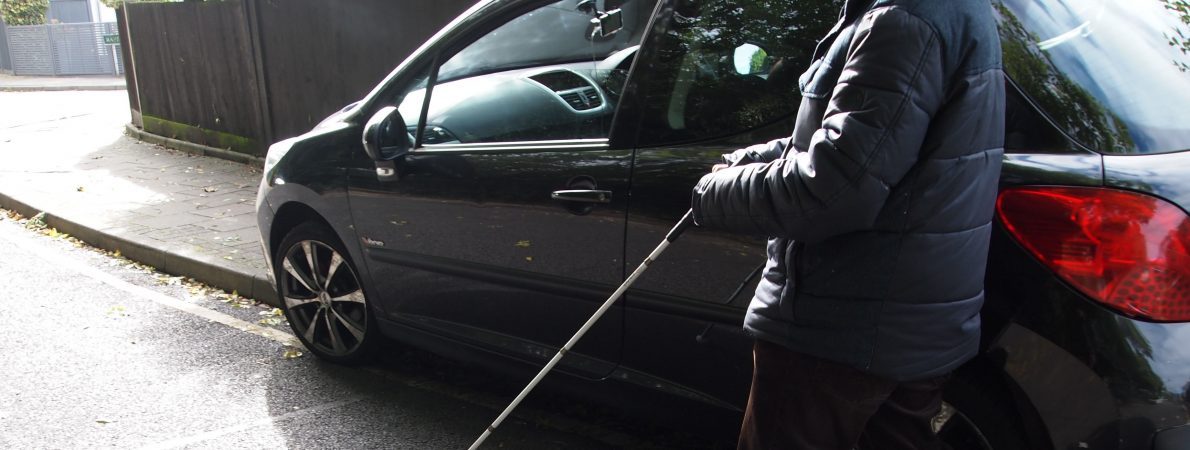 Black hatchback vehicle is parked with 2 of its wheels on the pavement, blocking pedestrian access close to a fence. A visually impaired person using a long cane is on the main road trying to navigate a way past the vehicle.