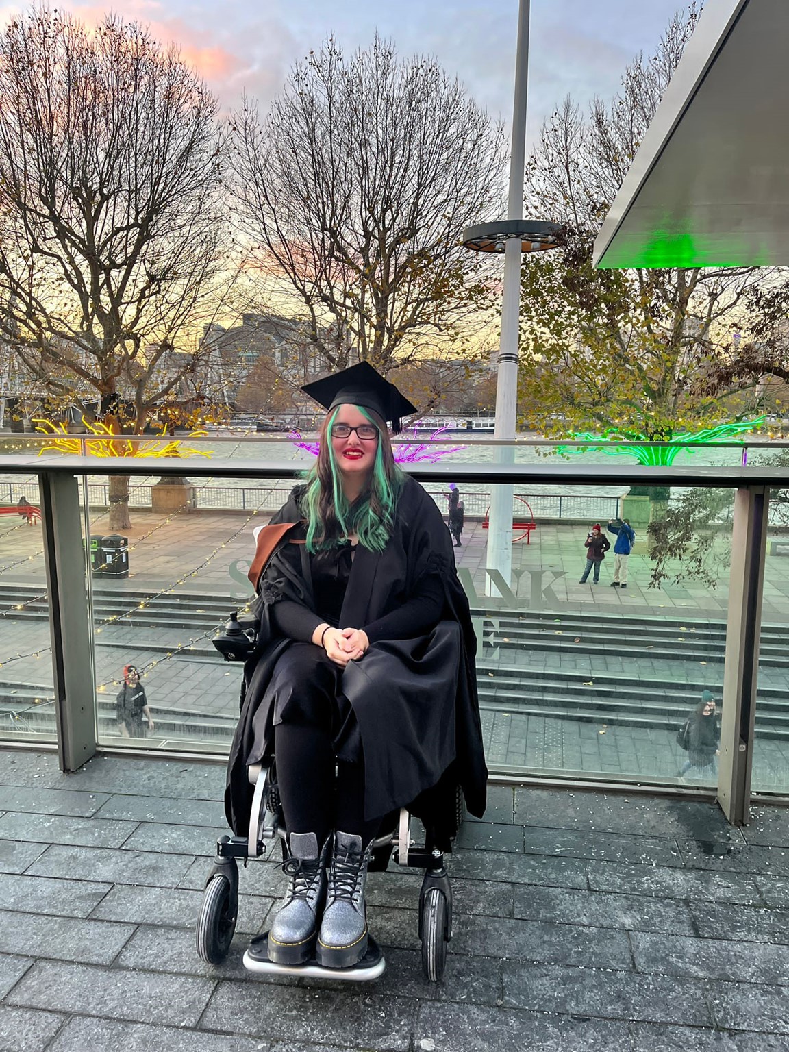 Rowan, a woman with green hair, sits in her wheelchair wearing a black graduation cap and gown. She is smiling at the camera. Behind her is a glass barrier and three trees lit up from their bases each with a different colour light of yellow, pink, and green. Beyond the trees is a river with stone university buildings on the opposite bank. The sun is setting.