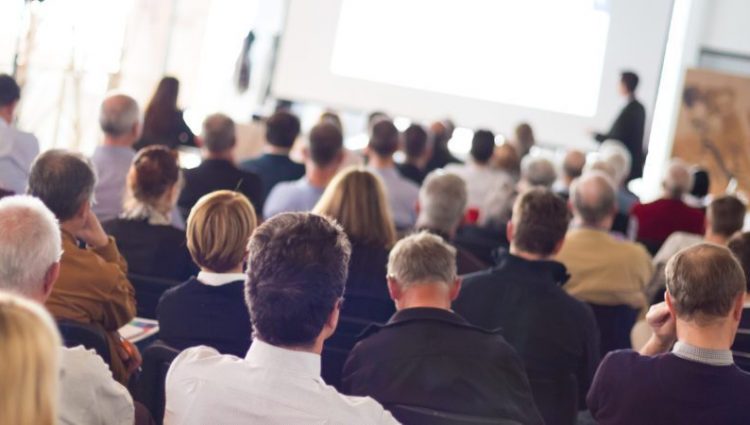 Photograph of an auditorium packed with people watching a presentation in the distance by a smartly dressed man standing in front of a screen. ages of the adult audience vary from young to old.