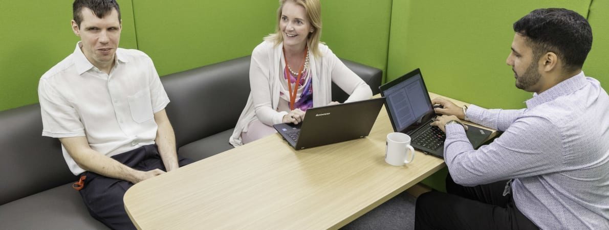 Three people sat in a row in a work environment. To the left is a man and to the right are a women and a man both using laptop computers.