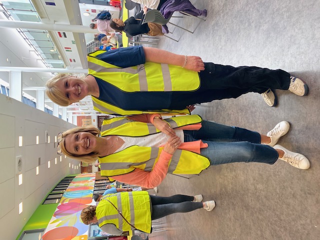 Photo shows John Lewis partners, Amanda McKenna and Jenny Slater wearing hi-vis jackets and stood in a sports centre. A female is walking past in the background.