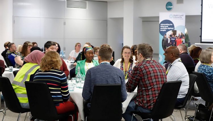 Sight Loss Councils' volunteers sat around table in a reception room. In the background is a Sight Loss Councils banner.