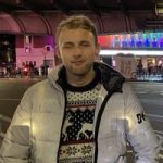 A head and shoulders picture of Aleks, a blonde man wearing a white jacket and a Christmas jumper, standing on the side of a busy road. He is looking at the camera.