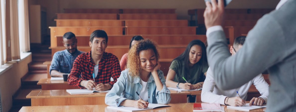 Group of students is listening to teacher and writing sitting at tables in classroom while male professor is talking and gesturing.