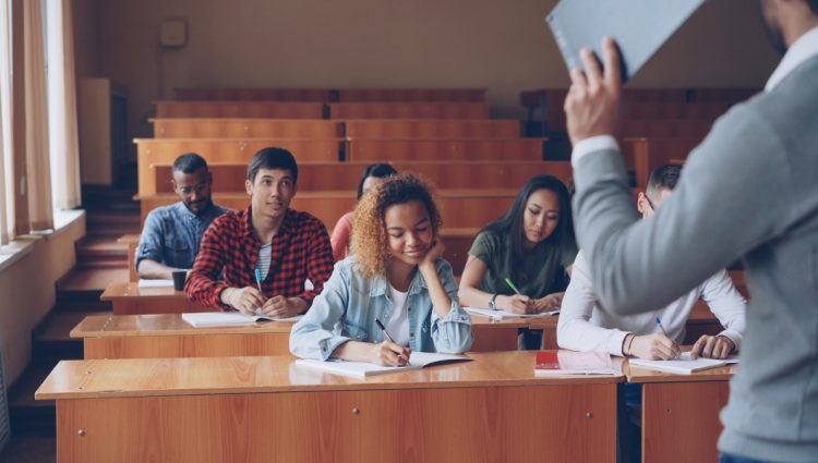 Group of students is listening to teacher and writing sitting at tables in classroom while male professor is talking and gesturing.