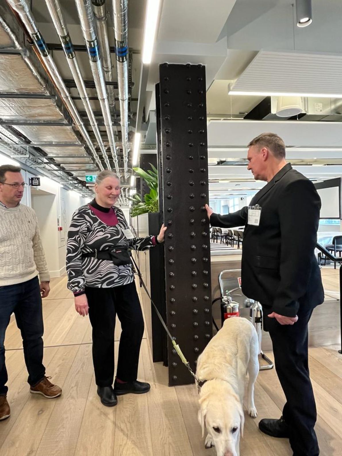 Image shows John (left) welcoming Johnathan, Mary and her guide dog Lewis (right) to the new PageGroup head office. They are all stood in a large open plan area, with a girder in between which Johnathan and Mary are touching with their hands