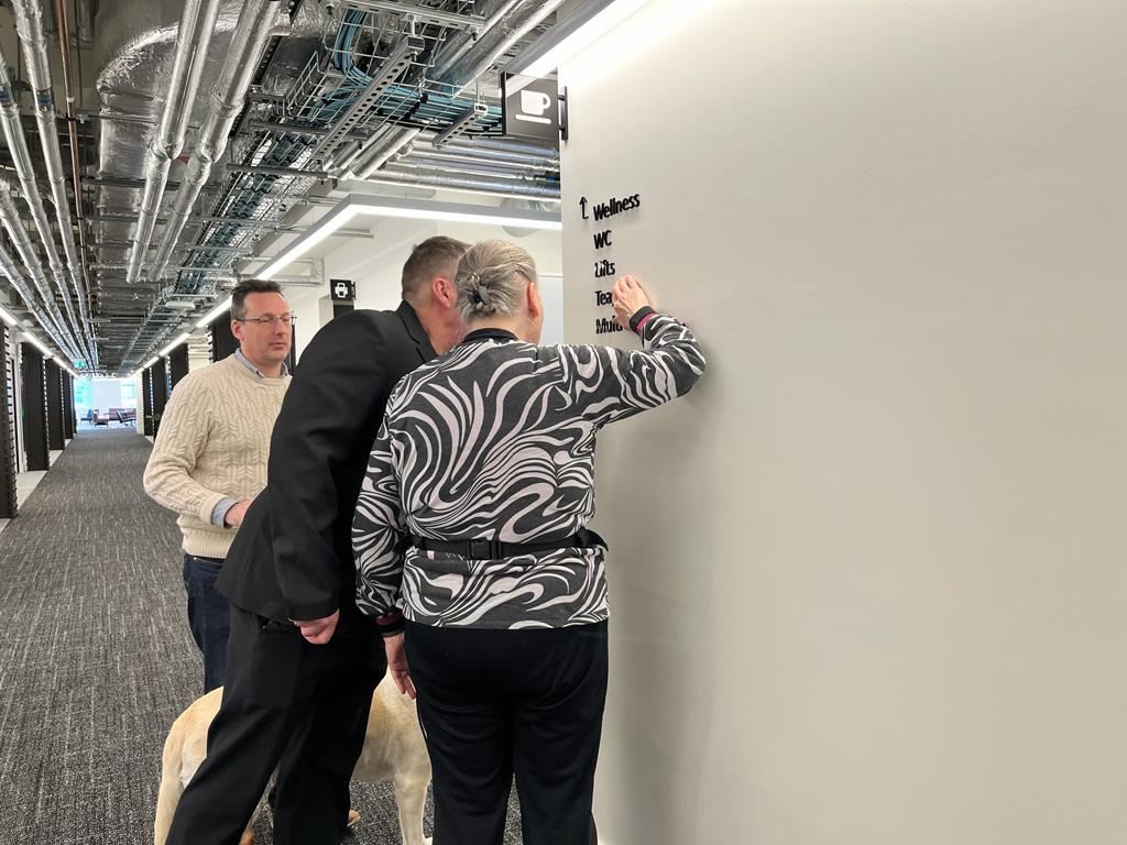 Image shows Mary touching the tactile signage as Jonathan inspects it, while John looks on.