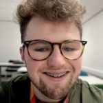 A head and shoulders picture of Tom, a young man with glasses, looking at the camera and smiling.