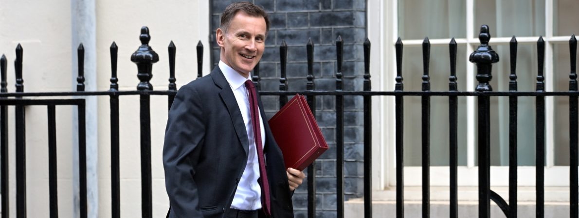 Chancellor Jeremy Hunt holding a red folder walking outside a building and looking at the camera.