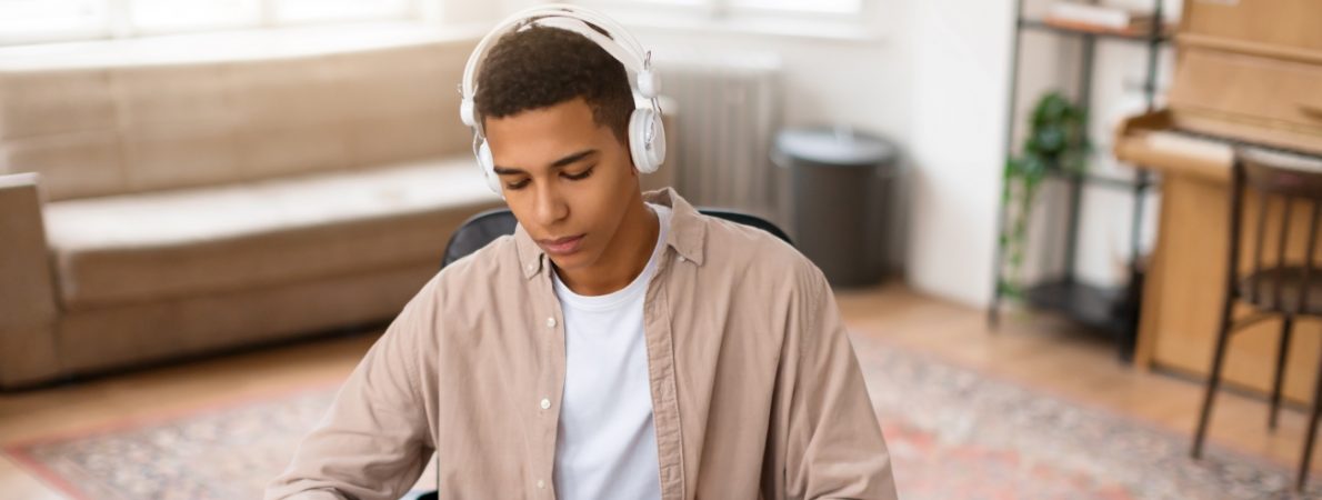 A focused young male with headphones studies from a book at a wooden desk with a laptop, coffee mug, and eyeglasses nearby.