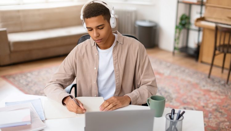 A focused young male with headphones studies from a book at a wooden desk with a laptop, coffee mug, and eyeglasses nearby.