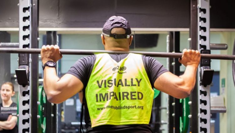 Image shows a gym goer in a squat rack at the gym. He has a barbell with weights on his back and is wearing a hi-vis vest which says 'Visually impaired'