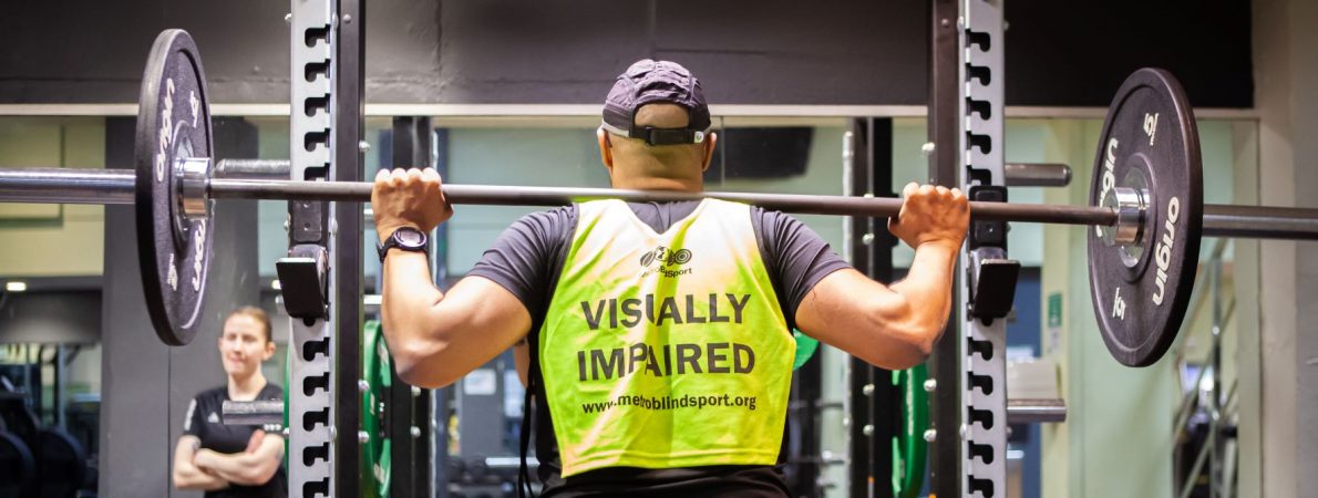 Image shows a gym goer in a squat rack at the gym. He has a barbell with weights on his back and is wearing a hi-vis vest which says 'Visually impaired'