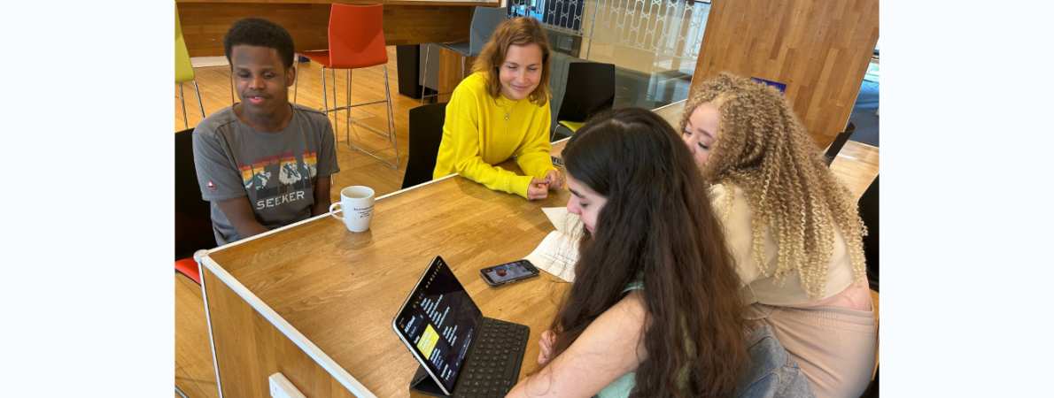 3 students sat around a table with Muller staff. They are making notes on an iPad with TPT offices in the background.