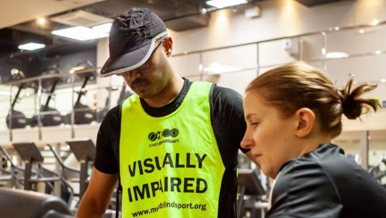 A woman showing a man with a vest that says 'Visually impaired' how to use the treadmill in a gym.
