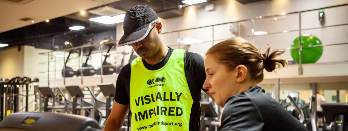 A woman showing a man with a vest that says 'Visually impaired' how to use the treadmill in a gym.