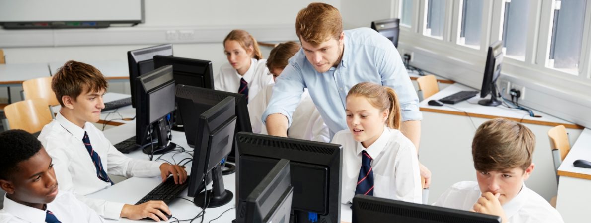 Teenage Students Wearing Uniforms Studying In IT Class. A man standing is helping one of them showing them something on the screen.