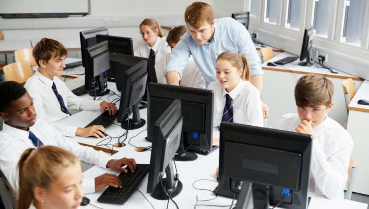 Teenage Students Wearing Uniforms Studying In IT Class. A man standing is helping one of them showing them something on the screen.