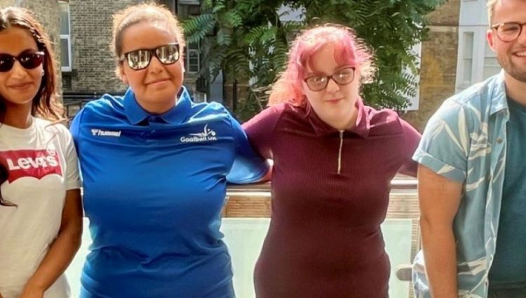 A picture of the Young Voices Sports team smiling at the camera, standing side by side in front of a glass railing with a building and trees in the background.