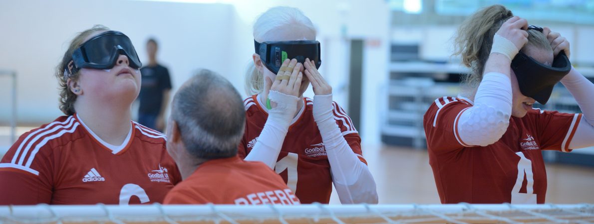 A close up of three goalball players wearing red kits, in various stages of putting their eye shades on.