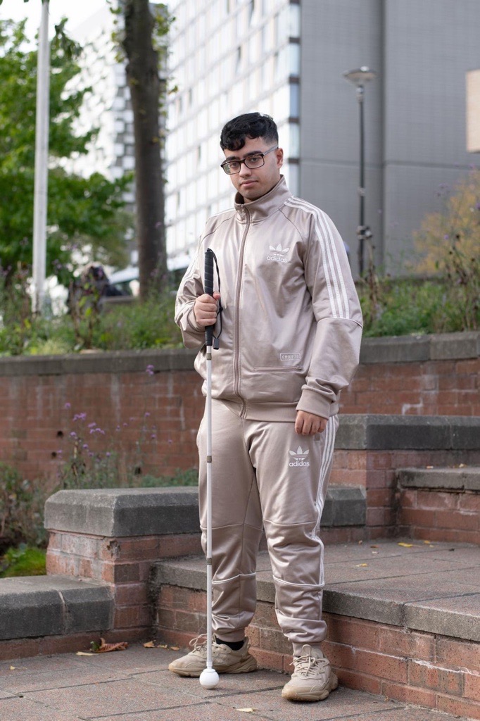 Picture shows Bilaal standing by some red brick stair outside of a block of apartments. He is holding a white cane while looking at the camera.
