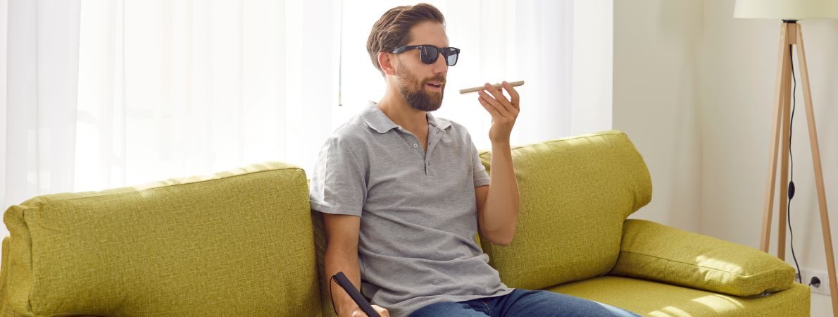 A blind man talks on the phone by holding the phone in front of his face. He is sat on a mustard coloured sofa in a minimalistic style living room.