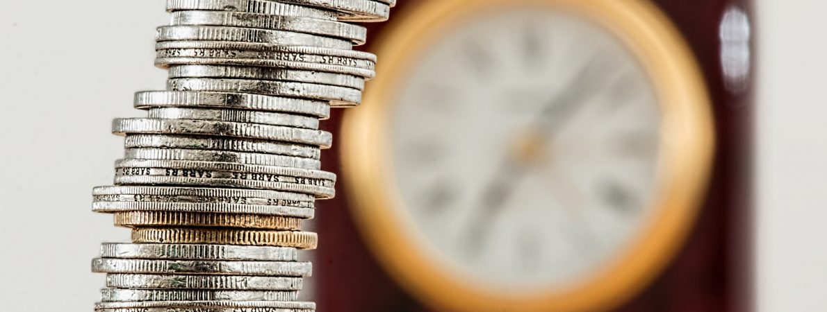 The picture shows a stack of various coins with a clock on the wall in the background