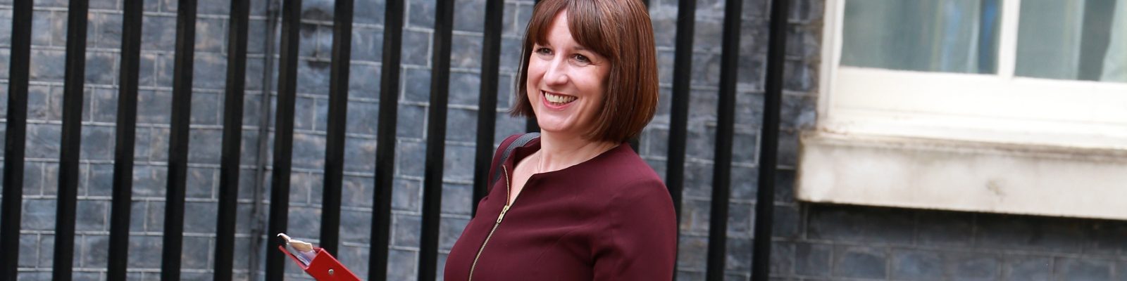 Chancellor Rachel Reeves holding a red folder walking outside a building and smiling.
