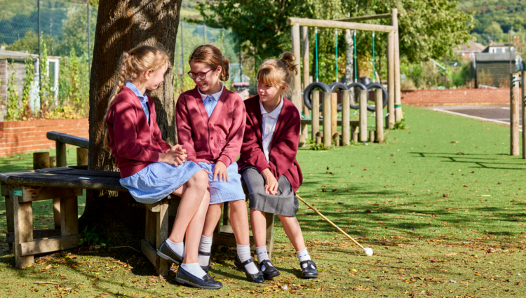 Three primary school aged children sit by a tree in their school uniform. Two are wearing a blue dress with a maroon cardigan over the top whilst the third is wearing a white blouse, grey skirt, the same maroon cardigan and is also holding a white cane. A playground is seen in the background.
