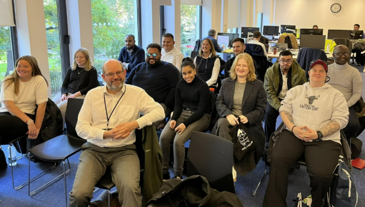Photo of latest of interns 2024 to 2025 cohort: A group of interns of mixed genders, age, and ethnicities sit in an open office environment with large windows that allow plenty of daylight.  