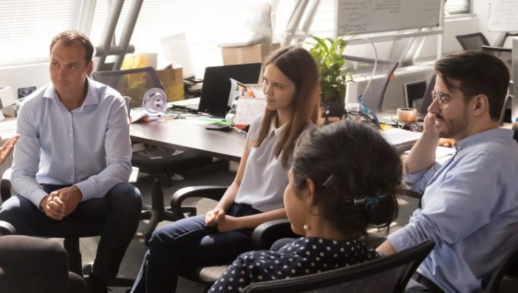 A group of people in smart office wear, discussing their ideas. They're sat in chairs arranged casually to form a loose circle.