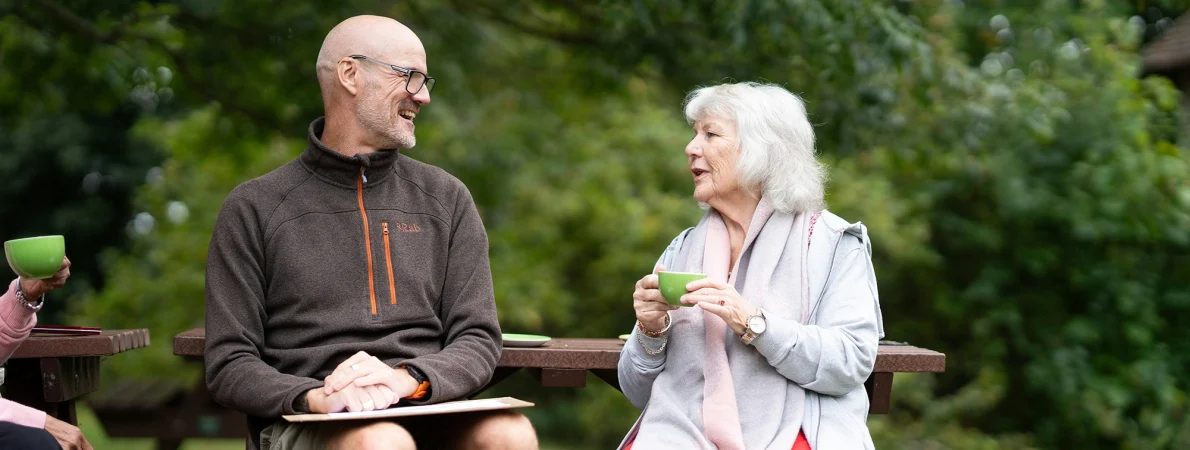 Two individuals chat on a park bench. One man is wearing shorts and an outdoors style fleece. He smiles as a lady with grey hair chats to him. She is holding a green cup of coffee.