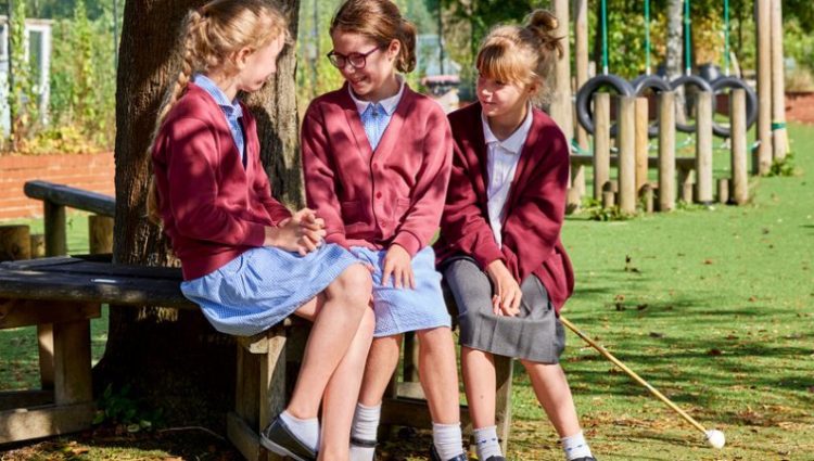 Three primary school aged children sit by a tree in their school uniform. Two are wearing a blue dress with a maroon cardigan over the top whilst the third is wearing a white blouse, grey skirt, the same maroon cardigan and is also holding a white cane. A playground is seen in the background.
