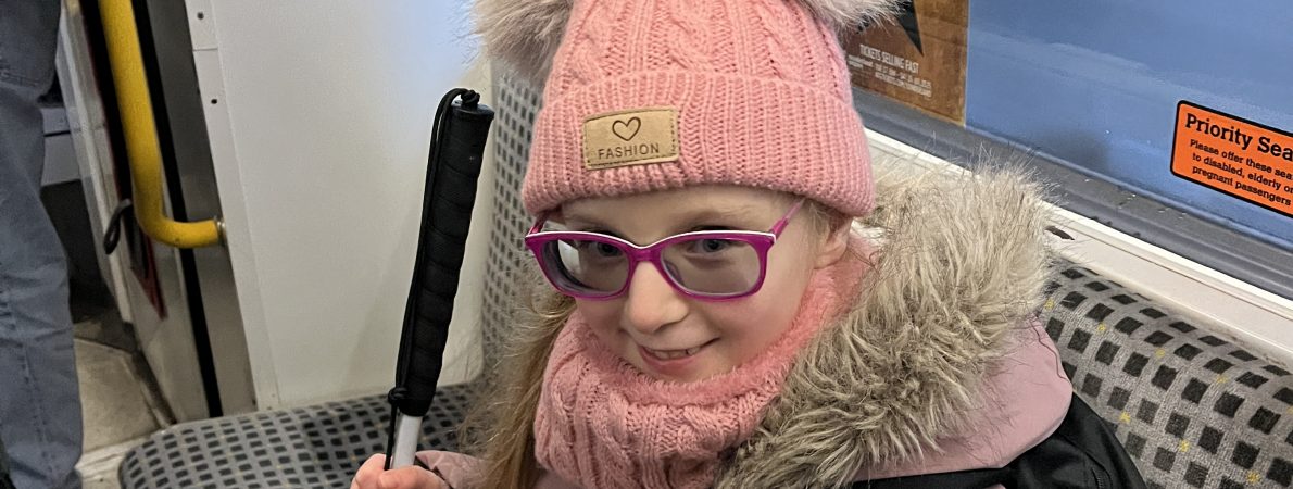 Lucy is sitting on a bus wearing a pink fluffy coat and matching hat, holding her cane and excited for an adventure.